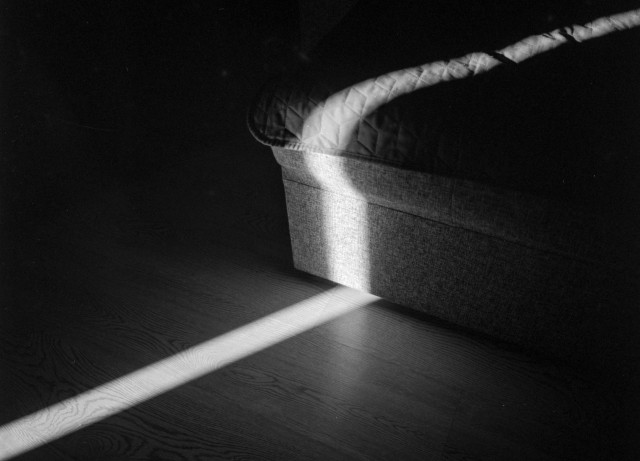 A diagonal beam of sunlight cuts across a dark room, illuminating the corner of a fabric-covered sofa and the wooden floor. The stark contrast between light and shadow creates a quiet, minimalist composition in black and white.