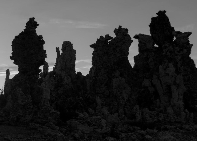 A high contrast black and white landscape photo of tufa towers which are mineral deposits that grow underwater into various rough sculpted shapes and as the lake level drops are exposed to the air. There are four distinct columns or towers standing side by side. Each is very rough with all sorts of angular shapes and jagged protrusions. They are very dark, being back lite by a bright sky.