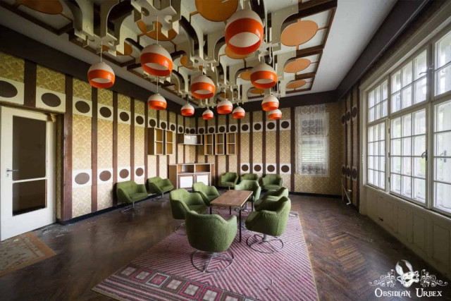 An abandoned, retro hotel room features olive green chairs, a patterned rug, and unique orange and white dome lights hanging from an elaborate ceiling