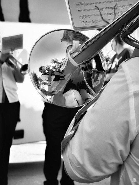La imagen en blanco y negro muestra a varios músicos tocando instrumentos de viento en lo que parece ser un evento al aire libre. En primer plano, se ve un trombón con una partitura sujeta en la parte superior, y en la campana del instrumento se reflejan los músicos y el entorno, incluyendo una persona con un ramo de flores. Al fondo, otros músicos aparecen ligeramente desenfocados.