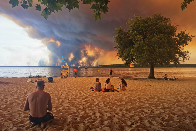 People sit on the beach of Moutchic as smoke rises into the sky from a wildfire. 