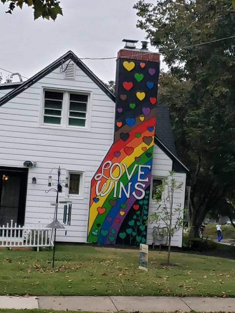 A two-story white house with a painted chimney. The chimney is painted with green hearts made to look like shamrocks at the bottom, a rainbow, across the middle, and  hearts in a variety of colors rising to the top. In white letters, across the rainbow, reads the phrase: Love Wins.