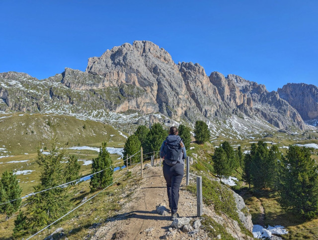 A person with a backpack hiking along a narrow dirt trail, viewed from behind. The path is lined with wooden posts and a rope fence, leading toward massive, rugged, partially snow-dusted mountains under a clear, bright blue sky.