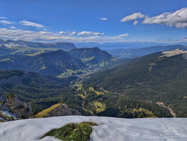 A high-altitude vantage point looking down into a vast, lush green valley nestled between dense forest-covered mountain slopes. In the foreground, a patch of snow sits on the ground, while the distant landscape features a small village and layers of hazy blue mountain ranges under a sky with scattered clouds.