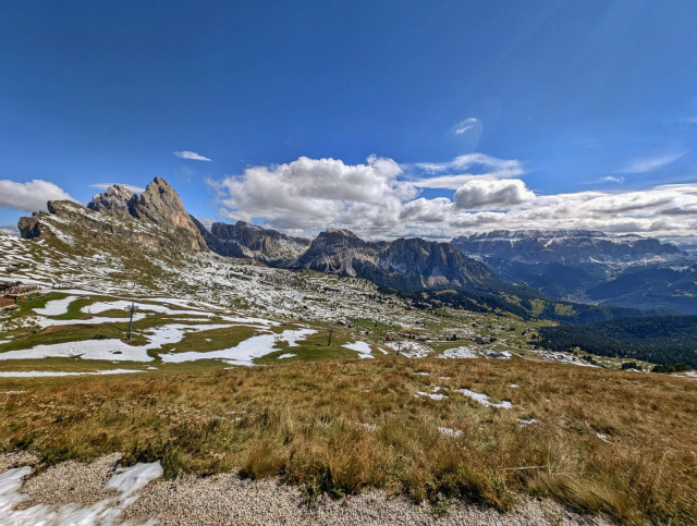 A sweeping panoramic view of a wide, high-altitude alpine meadow partially covered in patchy snow. The landscape is surrounded by dramatic, craggy mountain ranges under a vibrant blue sky with fluffy white clouds, with a few ski lift pylons visible in the distance.