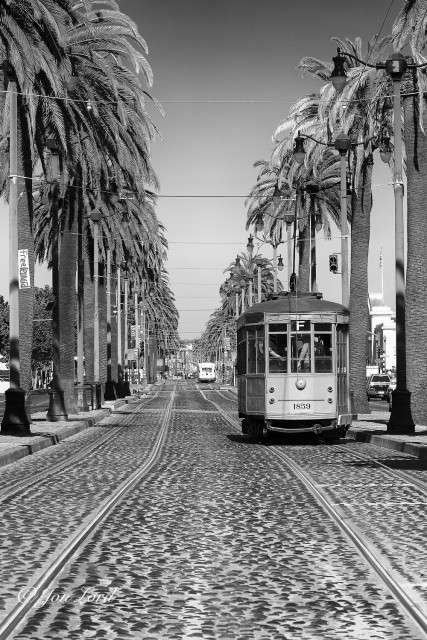 This is a black and white street photo in portrait format viewing along two long, straight and parallel sets of rail tracks with an old wooden Milanese tram (street car) on the right set of tracks. The Embacadero, San Francisco (2012).

In the foreground and stretching away from the camera and into the distance are the two sets of rail tracks. The camera is positioned between the two sets of tracks, about one metre above the surface and angled along the tracks. The rails are embedded into a bed of cemented medium sized, round stones: similar to those found on a beach. The two pairs of tracks are separated by a gap of about two metres. On either side of the tracks is a pavement area, lined with street lights and taller, six or seven metres tall, evenly spaced palm trees. Strung between the streetlights are cables stretched at right angles to the street that support the power cables that run above the rail tracks. On the right set of tracks is our old Milanese tram in motion away from the camera.  The tram is viewed from behind and at a distance of no more than ten metres. The tram is wooden above the wheels, has vertical, rectangular windows and is packed with people inside. Above the tram is a single gantry angled upwards and touching the power cable above. Ahead and above is a clear and cloudless sky, graduating from bright white at the base and becoming ever darker towards the top of the photo. 