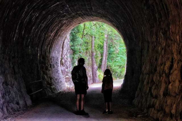 Two silhouetted figures—an adult with a backpack and a child—stand inside a dark, stone-built tunnel, facing the circular exit. The tunnel’s rough, textured walls and curved ceiling frame the scene. Beyond, a sunlit forest path with tall trees and dense greenery creates a dramatic contrast between the shadowed interior and the bright, wooded exterior.