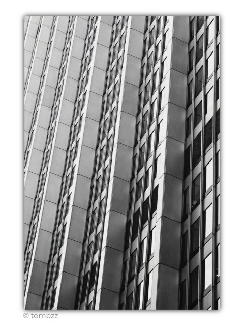 A black-and-white architectural photograph showing a tightly framed section of a modern building facade. Repeating vertical bands, rows of narrow windows and projecting modules create a raw, almost mechanical rhythm. The frame is slightly tilted, causing the regular lines to run diagonally and giving the static structure a distinct sense of movement. Light exterior panels contrast with dark windows, recessed sections and rectangular ventilation grilles. Soft light shapes the successive folds of the façade and emphasises their depth, while deeper shadows separate the individual segments. With no sky, street or clear point of reference, the building loses its scale and begins to resemble an abstract composition made of lines, rectangles, light and shadow. The image is defined by cool geometry, order and a consistent repetition of forms.