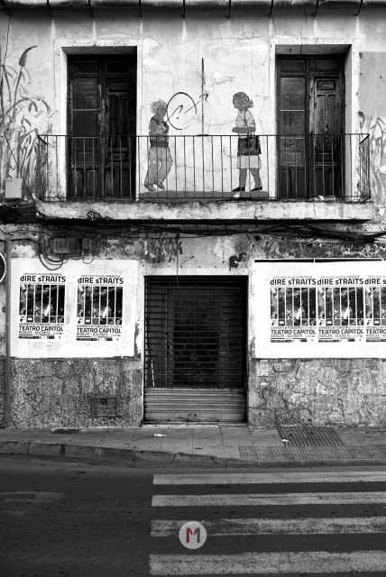 A vertical black and white photograph of a building facade showing an upper balcony and lower street level. On the top floor, two dark wooden doors flank a metal-railed balcony where a stencil-style mural depicts two figures facing each other near a circle. The bottom floor has a stone wall with a central barred window above a closed metal shuttered door, flanked by large posters that read: "WARNER MUSIC PRESENTA dire straits" and "TEATRO CAPITOL MADRID - ALCALÁ". In the foreground, a portion of a zebra crosswalk is visible on the street with a circular icon containing the letter "M" in one of its white stripes.