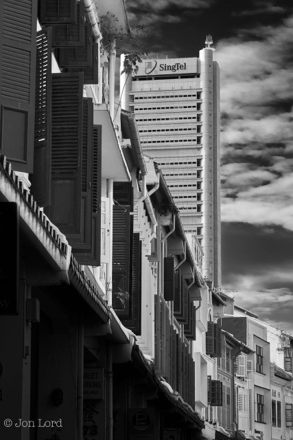 This is a cityscape photo in portrait format of a long row of historic, colonial-era terraced buildings in the foreground and a modern high-rise office block in the background. Amoy Street, Singapore (2012).

In the foreground is that row of old but restored colonial-era terraced buildings that start along the left edge of the photo and extend away from the camera for ten to fifteen metres before curving around to the right and then out of view. The location is a quiet and pleasant area of the city with mostly restored historic low- rise buildings, some residential, but mostly businesses and cafes. The camera is positioned about a metre to the right of the buildings (standing in the road) and aligned viewing along it's length. The buildings are a mix of two and three floors with the ground floor deep in shadow and mostly out of view with what is the pavement covered by the overhang from the first floor of the building, as was so common in that era to protect pedestrians from the tropical sun and even more tropical rain. All the windows on the one or two upper floors has vertical, rectangular wooden shutters, some open, some closed. Above the upper floor a small amount of sloping roof is visible with the guttering stretching along the edge of the tiles and with the metal down pipes extending down the front of the building. In the upper left corner are a few pieces of wild vegetation making a valiant effort to grow from the cracks and gaps. In the distance, a little less than one km away is a smart and modern silver grey, high-rise office block, probably around thirty floors tall with the words SingTel in large lettering together with a logo. Above and behind is a partly cloudy sky with scattered cirrus high clouds, the blue sky behind rendered to a jet black.
