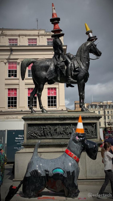 Vertical photo of the statue of the Duke of Wellington on a horse that stands outside the Gallery of Modern Art in Glasgow. The view is side on. It is customary for there to be a traffic cone on the duke's head, but today there are 4 stacked up, and his horse, which appears to have been turned into a unicorn with a sparkly horn, also wears cones (yellow and silver). At the foot of the statue is a temporary Scottie dog sculpture, also in profile, also wearing a cone on its head.