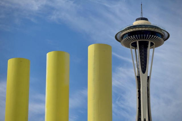 The image features three tall yellow cylindrical structures in the foreground, with the Space Needle in Seattle visible in the background against a blue sky with wispy clouds.