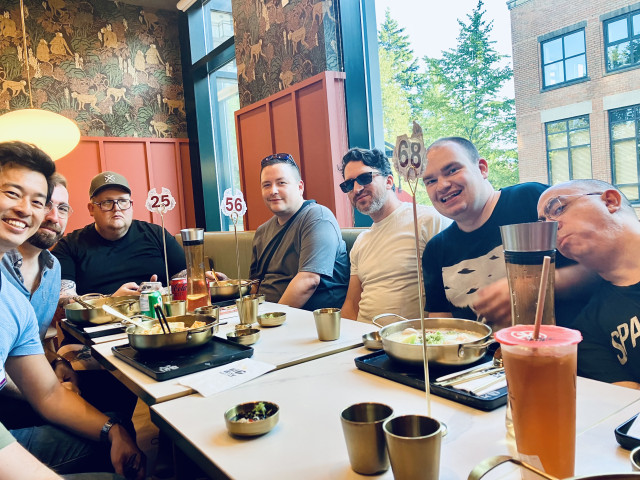 Seven men sit closely around a restaurant table, smiling and posing for a group photo over a Korean-style meal. Metal pots, bowls, cups, chopsticks, drinks, and tabletop burners cover the table, with numbered order signs rising above it. Large windows, coral-coloured paneling, and woodland-patterned wallpaper create a bright, lively backdrop.