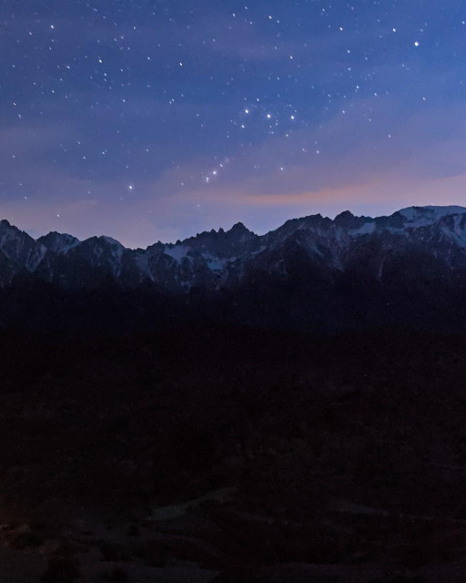 A late evening twilight photo of a snow covered mountain ridgeline with a star filled sky above and wispy waning light tinted orange sunset clouds above the mountain peaks. Below the mountain ridgeline is a very dark landscape with only the faintest outlines of features visible. A bright constellation is seen above the central peak. There are three bright stars along the top of the constellation and much dimmer ones that define somewhat of a box shape. In the lower left corner of the box several bright stars extend down at a 45 degree angle which are know as Orion's sword. 
