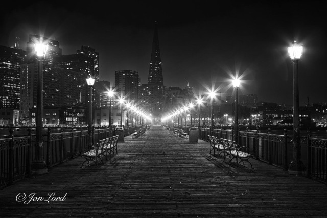 This is a grungy night time photo looking along a wooden pier aligned with and  towards the TransAmerica Pyramid in downtown San Francisco (2012).

Extending away from the camera and into the distance, about 150m away is the wooden pier. The rough wooden planks that form the surface cross at right angles to the pier and stretch away from the base of the image to the centre of the photo. On the side of the pier are metal railings with illuminated ornamental street lights spaced every 3 or 4 metres and casting a star like glow or aura around the lamp. Set inwards a metre of so from the railing are pairs of park benches facing outwards. The wooden planks, metal railings and street lights converge to a point with increasing distance towards the city end of the pier. Beyond is a night time cityscape with the high-rise buildings descending downwards from the business district on the left side to the low-rise residential buildings on the right. Aligned with the pier and in the middle ground, no more that one km away, is a tall, narrow, pyramid-like skyscraper with a sharp pointed top, lit with a single light. Like all of the surrounding high-rise buildings, some of the windows are lit, most are dark. Above is a ink black, dark sky, devoid of any detail. 

