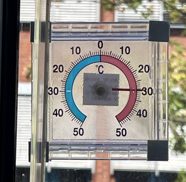 Nahaufnahme eines eine Fensterscheibe aufgeklebten Bimetall-Thermometers. Das Thermometer hat einen quadratischen, transparenten Körper mit einer runden Skala, die positive und negative Temperaturen bis 50 Grad Celsius darstellen kann. Aktuell zeigt es 30 Grad plus.