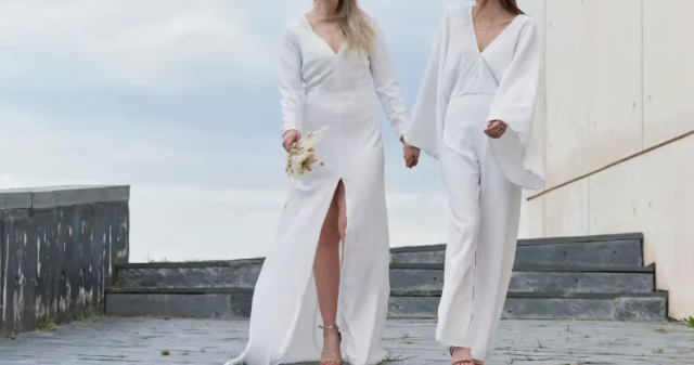 Two women in white bridal-style outfits walk hand in hand down outdoor steps. One wears a long dress with a thigh slit and carries a small bouquet, while the other wears a white pantsuit with wide sleeves.