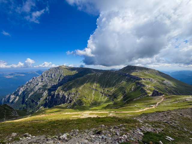 Wide panoramic shot of a vast mountain range under a bright blue sky filled with large, billowy white clouds. A dirt hiking trail winds across the grassy green ridge in the foreground, with steep rocky slopes cascading into the valley below. Farther along the ridge, small distant figures of hikers are visible, and a communications tower sits atop one of the distant peaks.