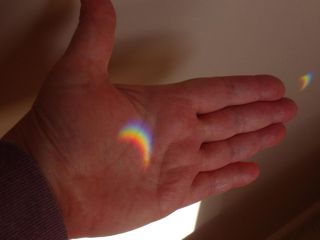 A colour photo of my left hand, held out with the palm facing the camera and the thumb up. Across the palm is a rainbow crescent of light, cast through a window on the opposite wall where a crystal is hanging. The partial solar eclipse has created the shape, it would usually be a bright circle.