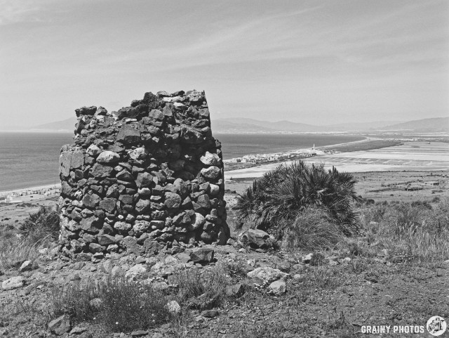The weathered ruins of a stone chimney stand atop a hill, overlooking a vast landscape. In the background, a tranquil seascape meets the horizon, with a small town and the Las Salinas Salt Flats visible along the coastline, surrounded by greenery and distant mountains in a black and white scene.