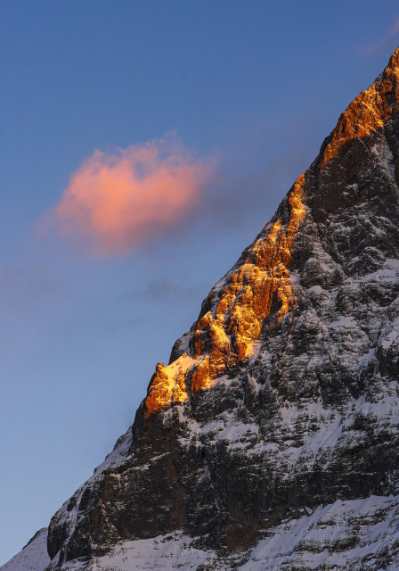 A snow-covered mountain peak illuminated by warm sunset light, with a soft pink cloud above against a blue sky.