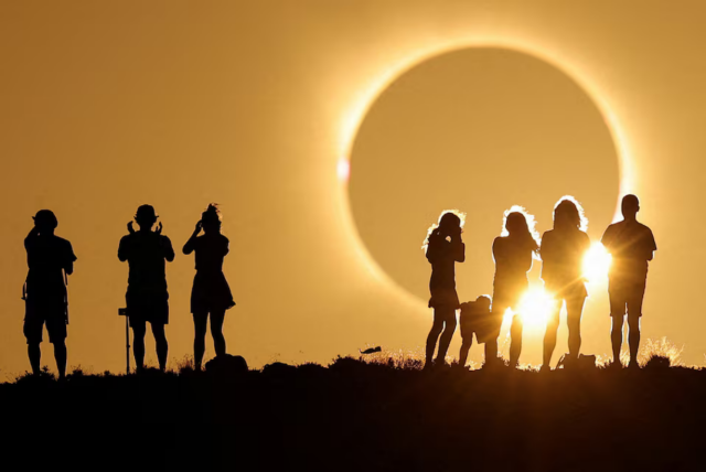 Silhouetted group of adults and children standing on a ridge against a warm golden sky, watching a dramatic total solar eclipse in Spain.

Detailed Alt Text
Visual Description:

A silhouetted group of eight people—a mix of adults and children—standing atop a dark hill or ridge against a warm, golden-orange sky. In the background, a dramatic total solar eclipse takes place, with the dark silhouette of the moon completely obscuring the sun, surrounded by a glowing rim of light and bright solar flare highlights along the horizon.

Left Side: Three adults stand in profile; two appear to be holding up cameras or solar viewing glasses to their faces.

Right Side: A group of five figures, including a young child leaning forward, stands silhouetted against a brilliant burst of sunlight breaking past the edge of the sky.

Lighting & Tone: Warm amber and golden tones fill the frame, capturing a striking moment of crowd awe during the eclipse.
