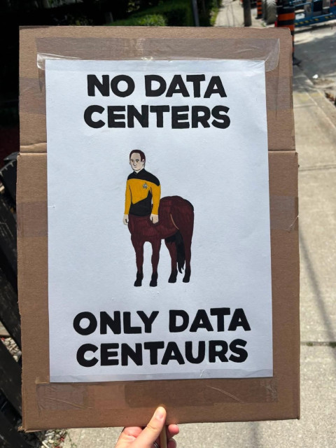 A hand holds a cardboard sign featuring a humorous illustration of a character in a Starfleet uniform with the text: "NO DATA CENTERS / ONLY DATA CENTAURS." The sign is placed outdoors, and the background shows a street.