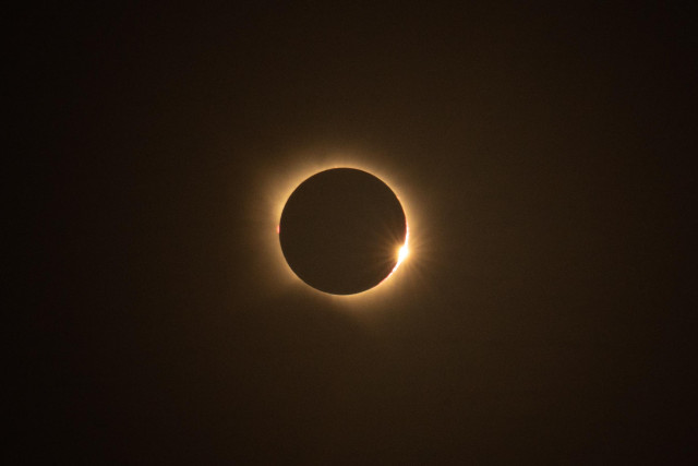 Photograph of a total solar eclipse. Against a dark, slightly brownish sky, the black disk of the Moon is visible, surrounded by the softly glowing solar corona. On the right side, the first intense ray of sunlight emerges from behind the Moon, creating the “diamond ring” effect.