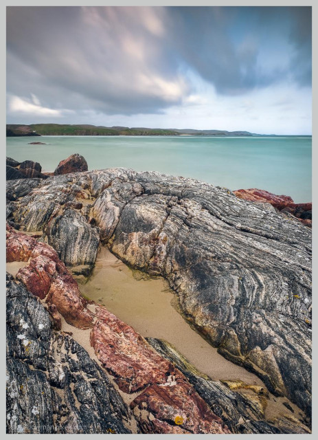 A photograph of Uig Bay on the Isle of Lewis in the Outer Hebrides of Scotland at low tide, showing a rugged shoreline covered with weathered, richly textured rocks, shallow pools of water and areas of pale sand. The rocky foreground leads towards a calm turquoise sea, with the distant coastline beneath a dark, brooding sky.