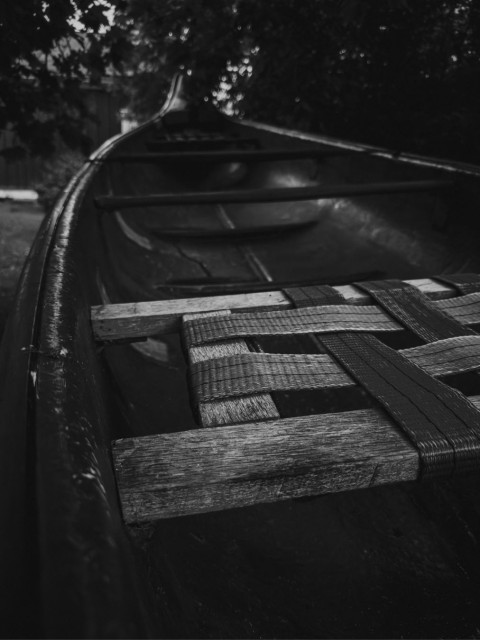 A black and white close-up of an old wooden canoe, focusing on its woven cane seat with worn wooden slats. The dark, glossy hull curves along the top of the frame, with blurred foliage visible in the background, suggesting a dim evening light.