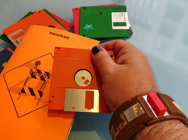 Photo of some stacks of multicolor 3.5 inch floppy disks and a HOPE 26 program. The hand of the Undersigned reaches in, holding a floppy. The Undersigned is wearing a "Blake's 7" teleport bracelet.