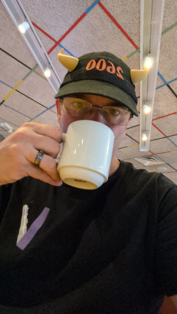 Middle aged IT guy in a 2600 hat with gold horns coming through and a Veilid T-shirt it is sipping a cup of coffee. 