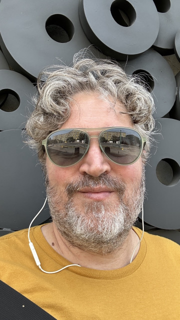 Before haircut. Scruffy beard, frizzy hair. I'm standing in front of a sidewalk art installation of black disks with center holes. I'm wearing wineglasses, white wired headphones, and a golden yellow T shirt.