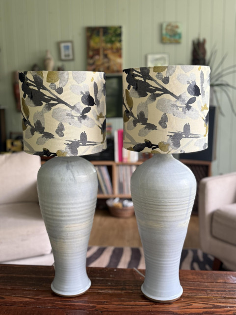 Two decorative lamps with floral-patterned shades sit on a wooden coffee table. The lamps have tall, rounded ceramic bases in muted blue tones, while the shades feature a watercolor design of gray and gold leaves on an off-white background. They sit on the coffee table in my living room with furniture, art work and a shelf of vinyl records blurred in the background. 