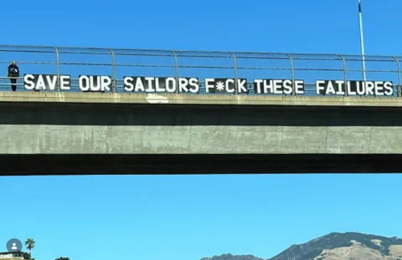 Against a backdrop of blue sky and mountains, California bridge protestors have put up letters on a bridge saying "SAVE OUR SAILORS - F*CK THESE FAILURES"
