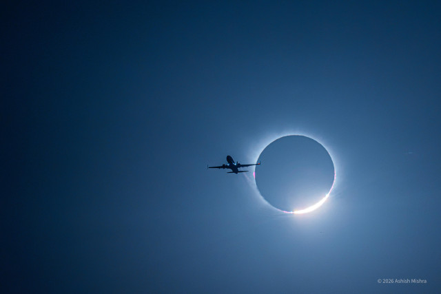 A passenger aircraft flies past a solar eclipse. Its dark silhouette is captured against a deep blue sky as a thin glowing ring of sunlight and bright flare shine around the covered sun behind it.