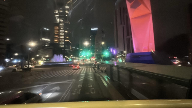 Night view from front seat upper deck of metrobús 7 traveling down the Paseo de la Reforma in Mexico City. A large pink lighted sculpture looms at right. A fountain is in the center of a circle at left. 