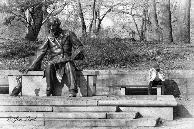 This is a black and white street photo in landscape format of a scene in a city park with a large statue of Charles Dickens sitting on a park bench and feeding a duck while to the right is a live man sitting on another park bench looking at the ground. Central Park NYC (2012).

In the foreground are a few pale and wide steps leading up to a stone plinth. On the plinth is the statue of Dickens sitting on a oversized park bench. The statue is dark, about three metres tall, fills a little over half the height of the photo and is viewed from about five metres away. On Dickens left leg rests a large open book. His right arm is stretched out and downwards towards a large statue of a duck as if to feed or stroke it. To the right of Dickens and the duck is a normal sized park bench with a man seated on the right side. He is wearing a baseball cap, white sweat-shirt and dark trousers. He is looking downwards and possible talking on the phone. In the immediate background is a bank rising upwards a few metres with multiple, randomly scattered trees extending across and behind. In the far background, about one km away, are numerous mid-rise apartment buildings. 
