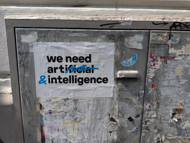 A3 großes Plakat dass auf einer Plakat- und Stickerresten übersäten Stahltür klebt. In dicken schwarzen Druckbuchstaben steht "we need artificial intelligence" - allerdings ist mit blau der hintere teil von "artificial" durchgestrichen und ein & dahinter gesetzt, womit sich "we need art & intelligence" ergibt. 