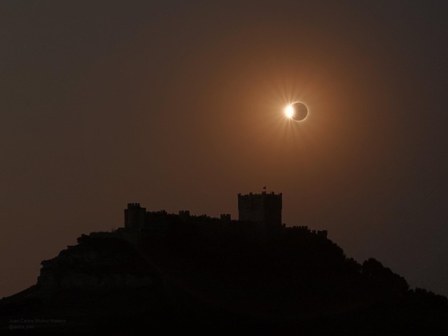 A photograph of a castle silhouetted against an orange sky. Above the castle, the Sun appears as a bright ring around a dark core. The Moon isn't completely blocking the Sun, though, and some sunlight shines brightly through the left side, like a diamond ring.