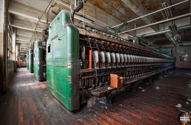 Rows of old textile machines with yarn and spindles still in place. A long wood bench is in front of the most prominent row. The floors are wood and pipes run overhead along the ceiling. The machines are a bright green.