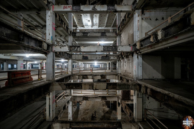 View of a cavernous interior; through a gaping hole in the floor the floors above and below are visible. The building has been stripped to bare concrete and the floors, walls, and support columns. Work lights placed sporadically about light the area