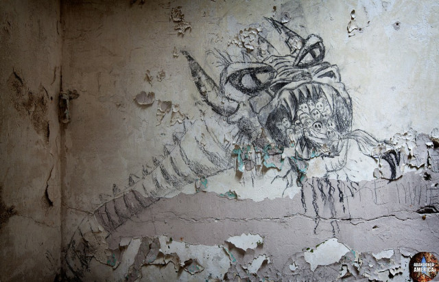 A decaying cell wall with peeling off white paint. There is a drawing on the wall of a serpentine monster with snake eyes, horns, and an open mouth with sharp fangs. Inside the mouth are circular white faces, barely detailed except for the eyes, staring out