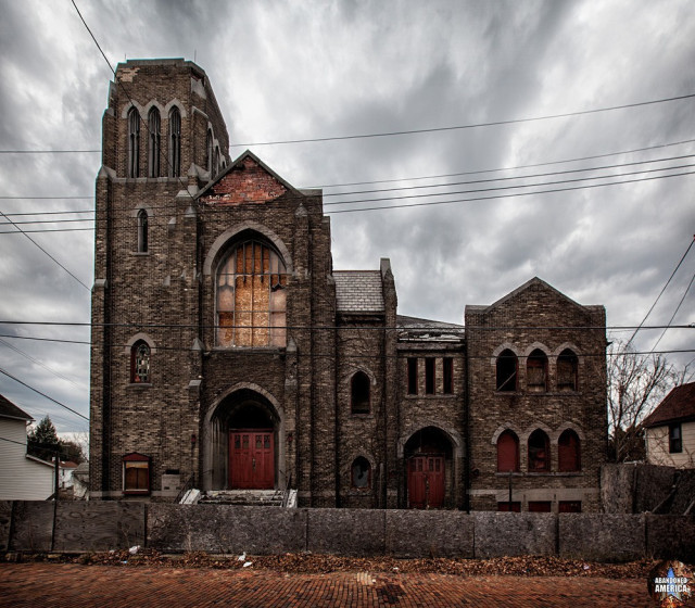 An exterior of a crumbling brownstone (?) church on a gloomy day. The windows are boarded or broken, and the doors and shutters are red.