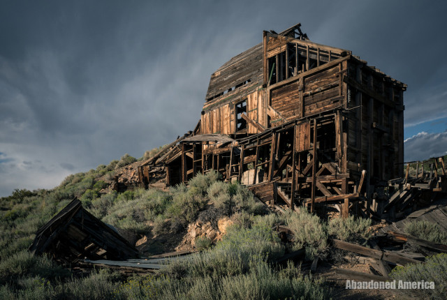 Beneath a moody dark blue sky, a collapsing wooden building approximately 3 stories tall sits on the side of an embankment. Stairs and walls are visible but it looks like a strong gust of wind will knock it over. 