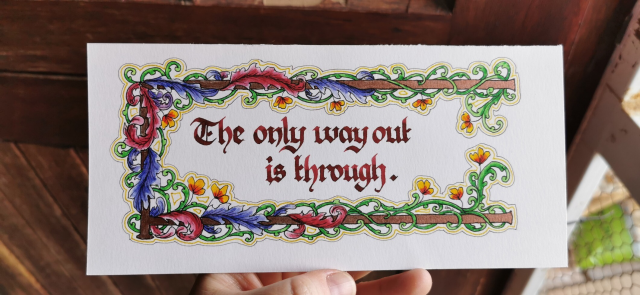 A calligraphy example, one short phrase on a narrow piece of paper surrounded by a gold bar with wrapped leaves and vines