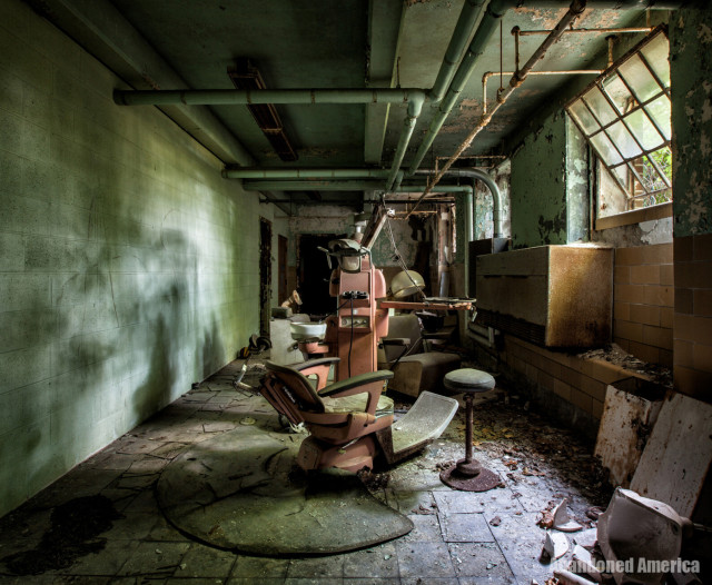 A long corridor with concrete walls and pipes running along the ceiling. Window wells on the right allow in light casting long shadows across the darkened room. In the foreground there is an antique pink dental chair, stool, and a corresponding piece of equipment with a light and spittoon. In the background a salon chair is visible.