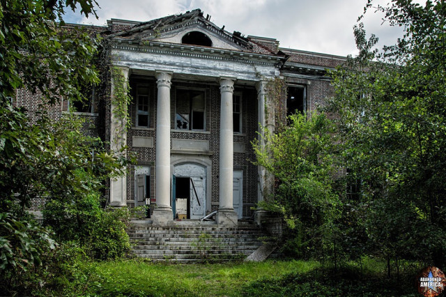 A ruined two story Neoclassical facade of an abandoned school. Steps lead up to columns that flank an open front door. Trees shroud the exterior.