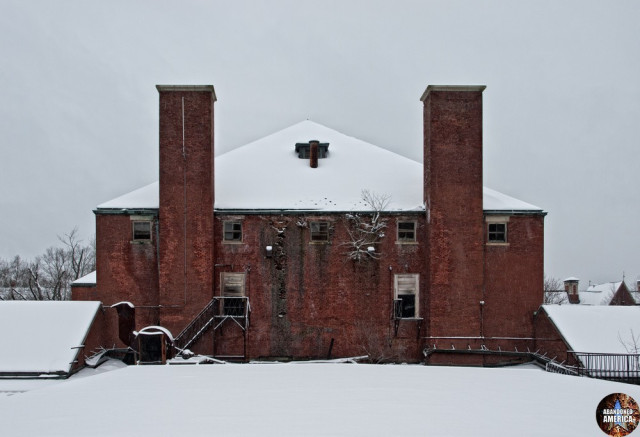Under a pale sky, the snowy forms of rooftops are visible. Ahead of the viewer a two story red brick segment with large chimneys rises out of the whiteness. Two doorways are on the side, one accessible via a metal staircase and the other broken