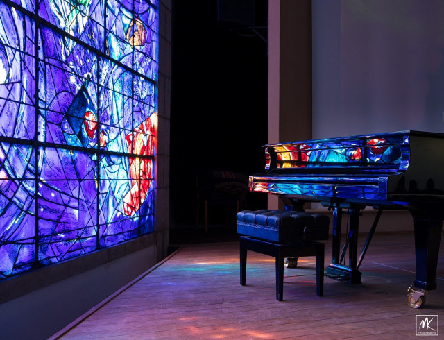 Color photo of the lower part of a large stained glass window with deep purples, blues and some reds designed by the artist Marc Chagall. Near the window is a glossy black piano reflecting the colors of the stained glass. 